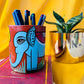Six coloured pens organized in a terracotta pen stand hand painted with a painting of a blue elephant placed in front of a plant pot