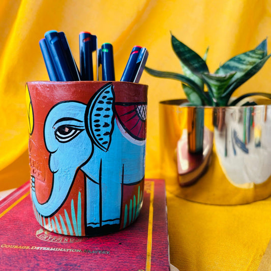 Six coloured pens organized in a terracotta pen stand hand painted with a painting of a blue elephant placed in front of a plant pot