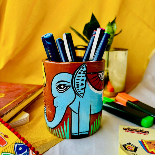 Six coloured pens organised in a terracotta pen stand hand painted with a motif of a blue elephant placed against a yellow background along with green and orange highlighter, sticky notes, a book and a plant pot.