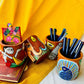 Two terracotta pen stands holding six coloured pens each hand-painted with peacock and musical instruments, one elephant-painted terracotta pen stand and a terracotta pen holder painted with a motif of tribal man is placed against a yellow background.