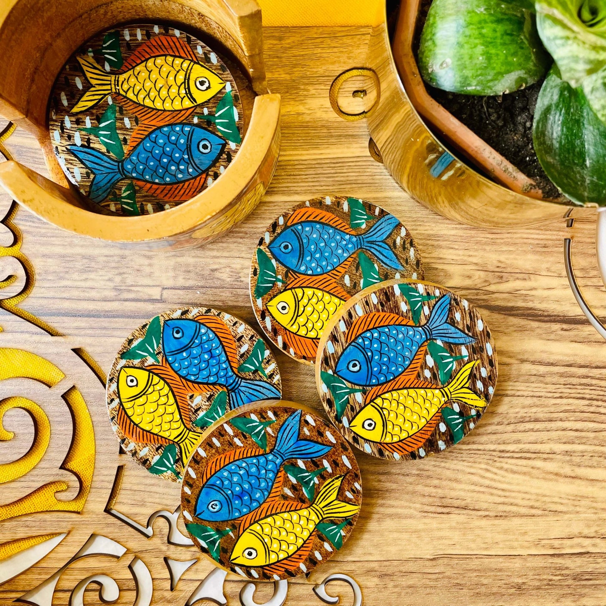 One pure mango wood round wooden coaster is placed in a wood coaster holder while 4 round wooden coaster hand painted with blue and yellow fish being displayed near a flower pot.