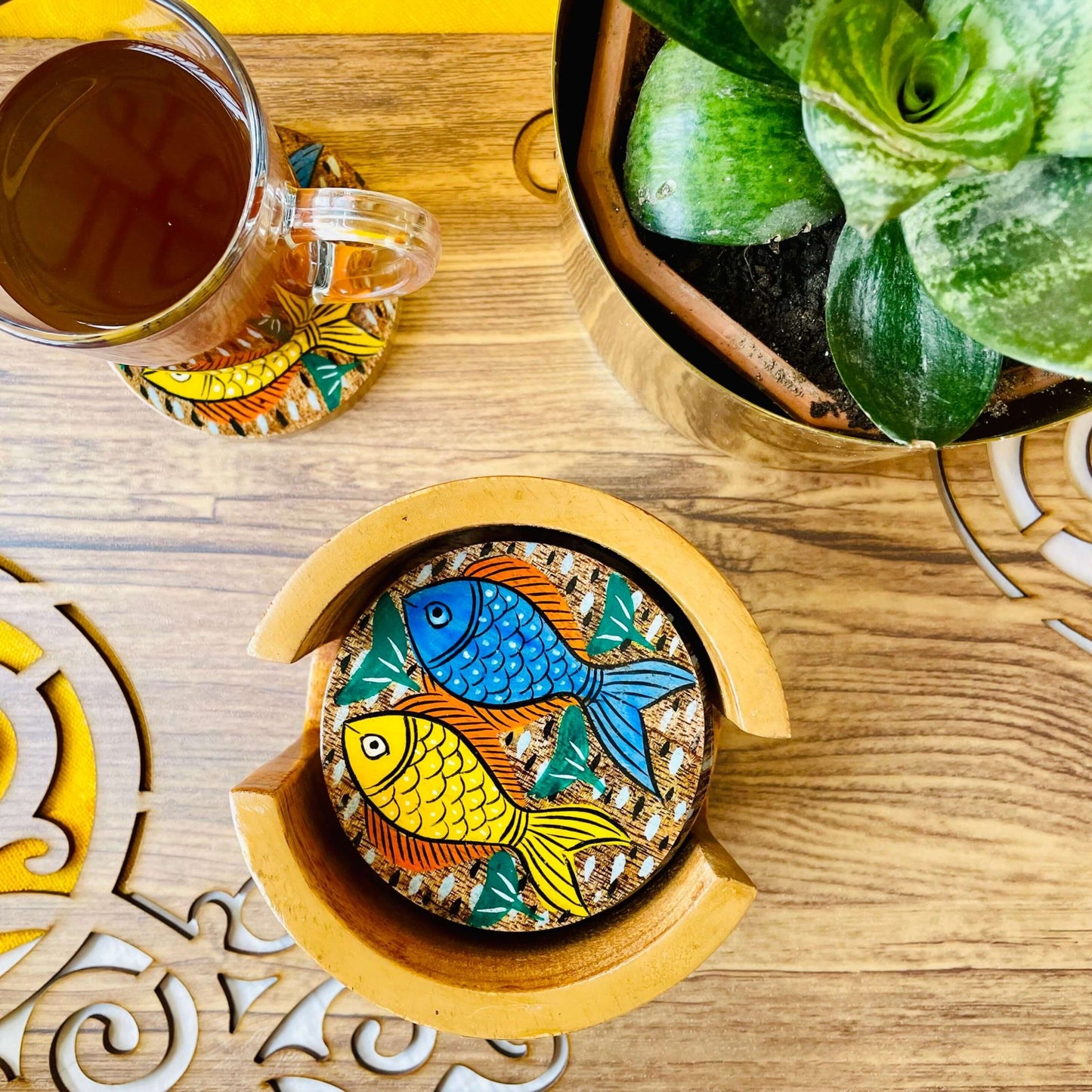 blue and yellow fish print round wood coasters placed in a round wooden coaster holder near a flower pot and tea cup placed on a round wooden coaster.