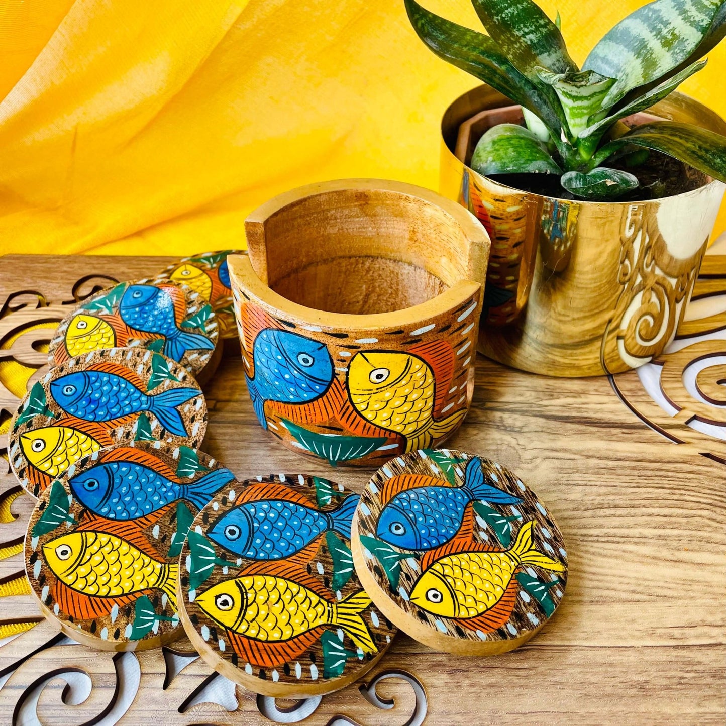 An empty coaster holder placed in front of a plant pot along with 6 round wooden coasters, all hand painted with blue and yellow fish motifs.