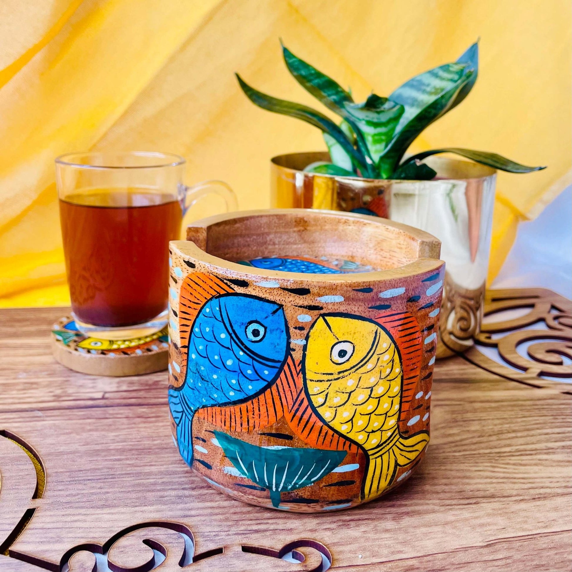 pure mango wood round wooden coaster holder with blue and yellow fish painted on it is being displayed with a warm cup of black tea and plant pot in the background.