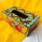 An aerial view of a 9”x5”x3” pure mango wood wooden tissue holder, handcrafted by rural artisans and painted with two red and yellow birds, tree branches and leaves motifs