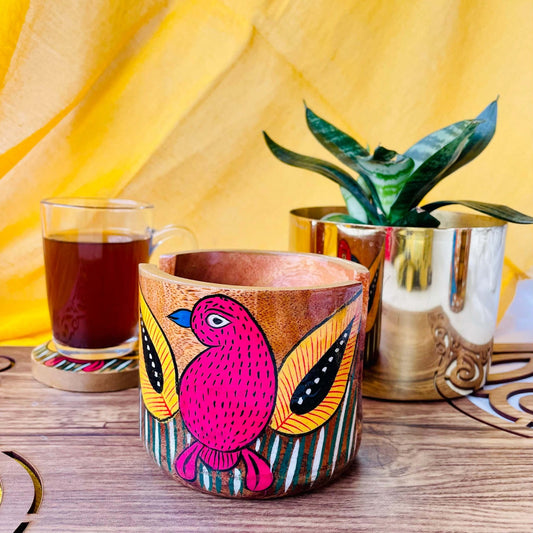 pure mango wood round wooden coaster holder with red birds with yellow feathers and blue beaks painted on it is being displayed with a warm cup of black tea and plant pot in the background.