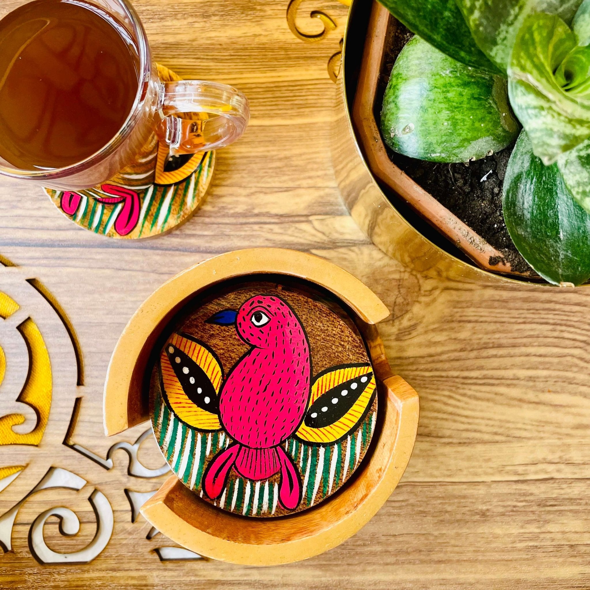 red bird print round wooden coaster placed in a round wooden coaster holder near a flower pot and tea cup placed on a round wooden coaster.