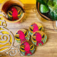 One pure mango wood round wooden coaster is placed in a wood coaster holder while 4 round wooden coasters painted with red birds having yellow feathers and blue beaks being displayed near a flower pot.