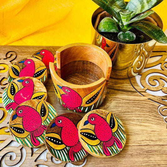 An empty coaster holder placed in front of a plant pot along with 6 round wooden coasters, all hand-painted with red birds with yellow feathers and blue beak motifs.