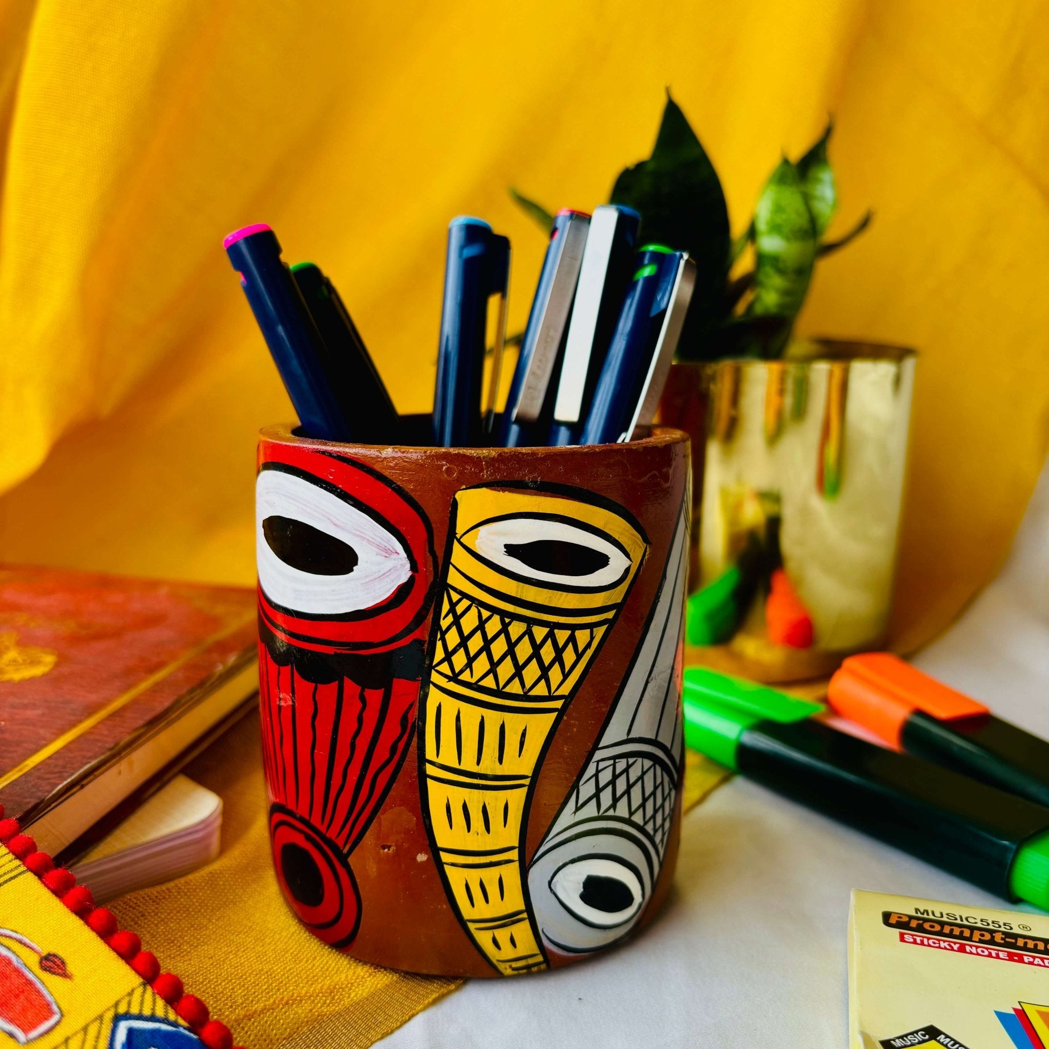 Six coloured pens organised in a terracotta pen stand hand painted with a motif of musical instruments are placed against a yellow background along with green and orange highlighter, sticky notes, a book and a plant pot.