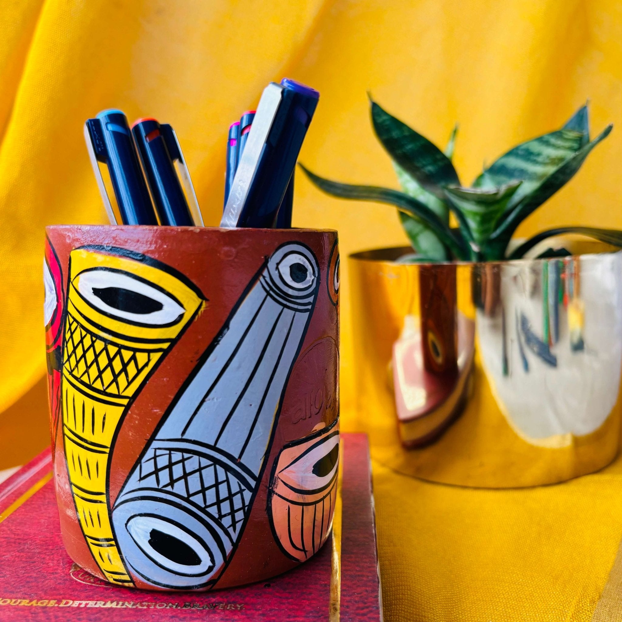 Six coloured pens organized in a terracotta pen stand hand painted with a painting of musical instruments placed in front of a plant pot