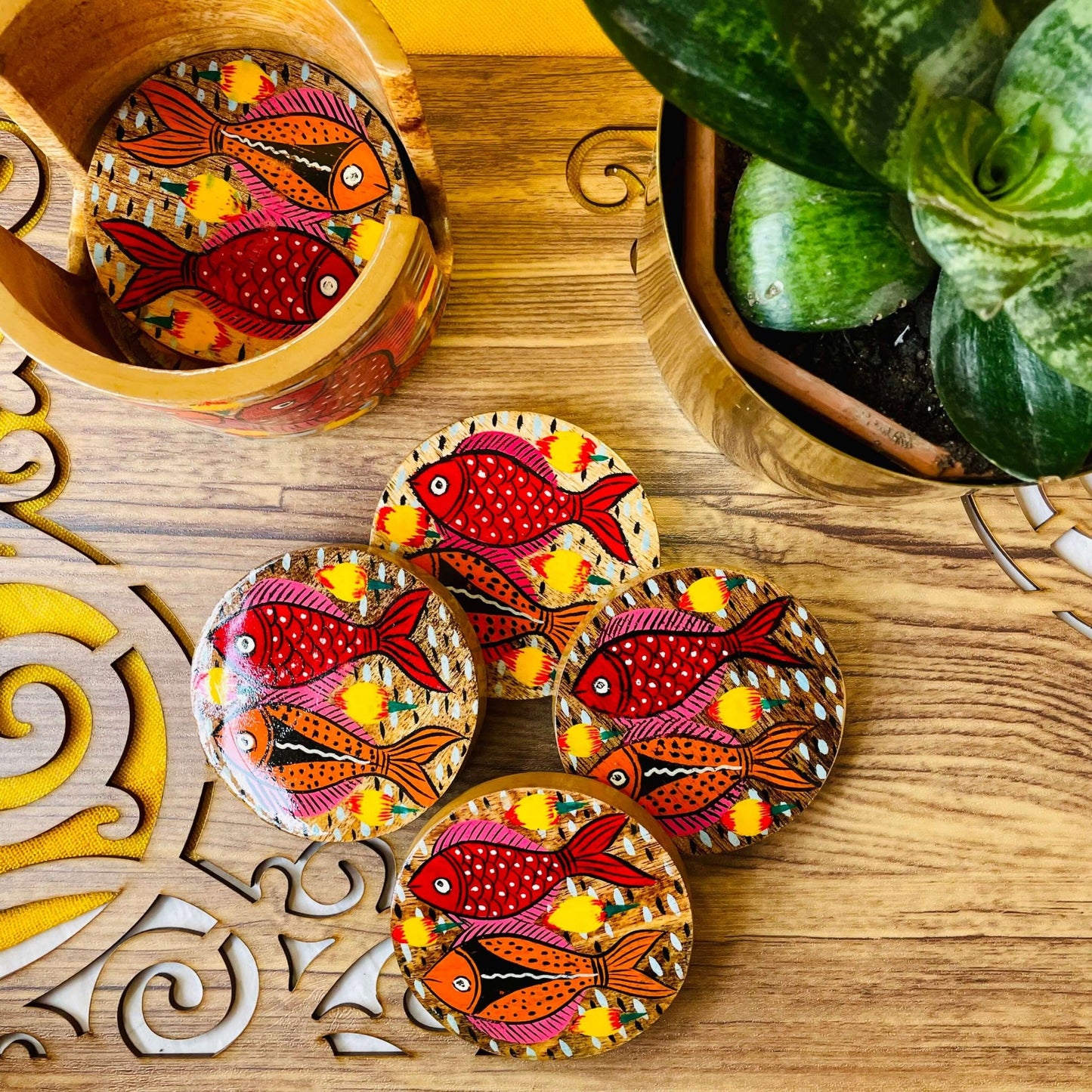 One pure mango wood round wooden coaster is placed in a wood coaster holder while 4 round wooden coasters hand-painted with red and orange fishes having pink and yellow fins are displayed near a flower pot.
