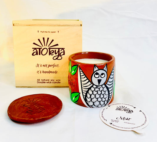 100% natural soy wax scented candle in a terracotta jar that is hand-painted with a grey owl motif. A seed paper candle dust cover is placed near the scented candle and has a wooden candle box and candle snuffer in the background.