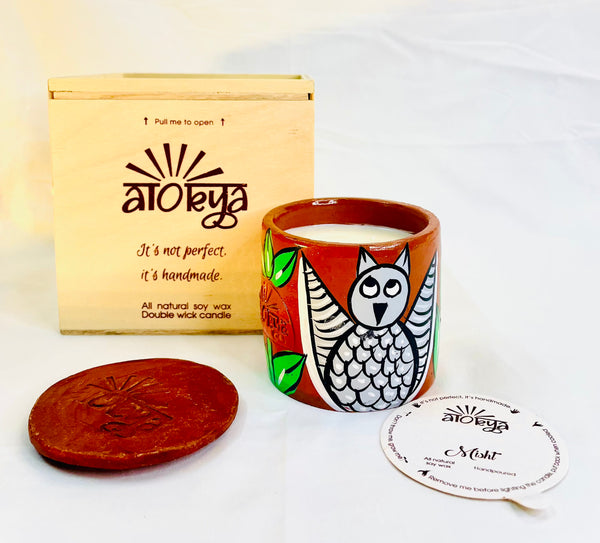 100% natural soy wax scented candle in a terracotta jar that is hand-painted with a grey owl motif. A seed paper candle dust cover is placed near the scented candle and has a wooden candle box and candle snuffer in the background.