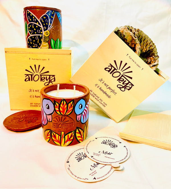 Two wooden boxes placed in the background with one having a 100% natural soy wax scented candle hand-painted with a yellow butterfly having blue wings motif on top of it and the other having a honeycomb comb paper in it. One 100% natural soy wax scented candle hand-painted with blue-coloured birds and yellow leaves around it is placed in the foreground along with a candle snuffer and two seed paper dust covers.