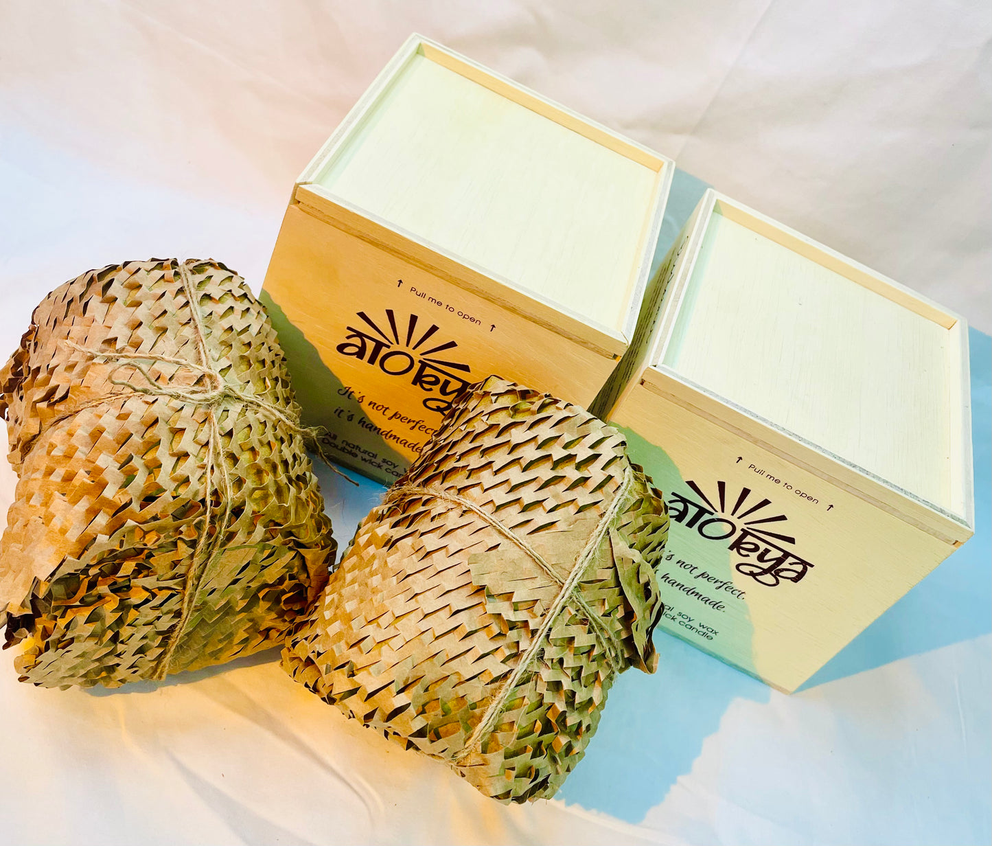 Two 100% natural soy wax scented candles wrapped in honeycomb paper are placed near two brown wood boxes.
