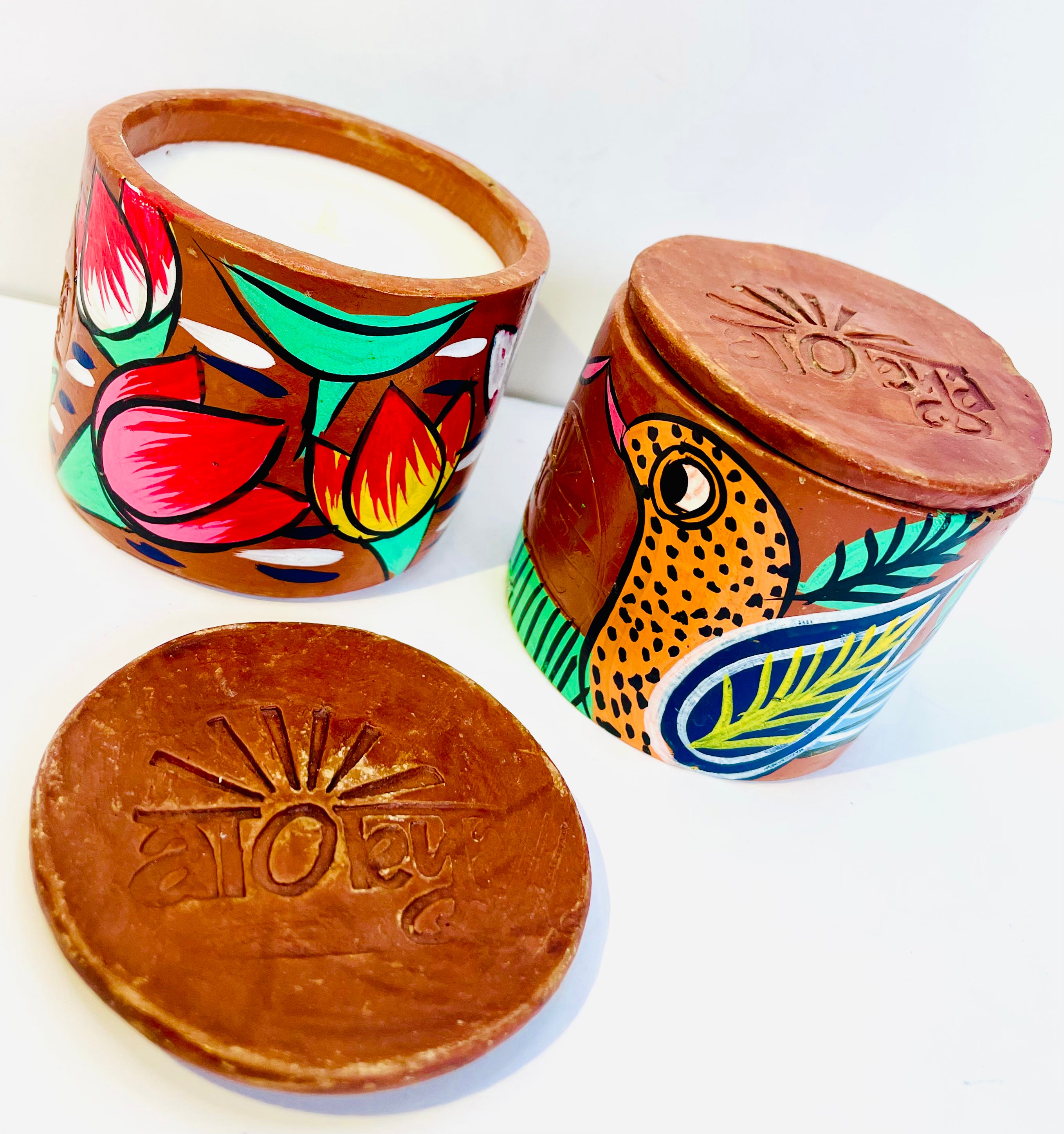 Two 100% natural soy wax scented candles, one hand-painted with an orange bird having blue and yellow wings and the other with pink lotus and green leaves are placed near a terracotta clay candle snuffer