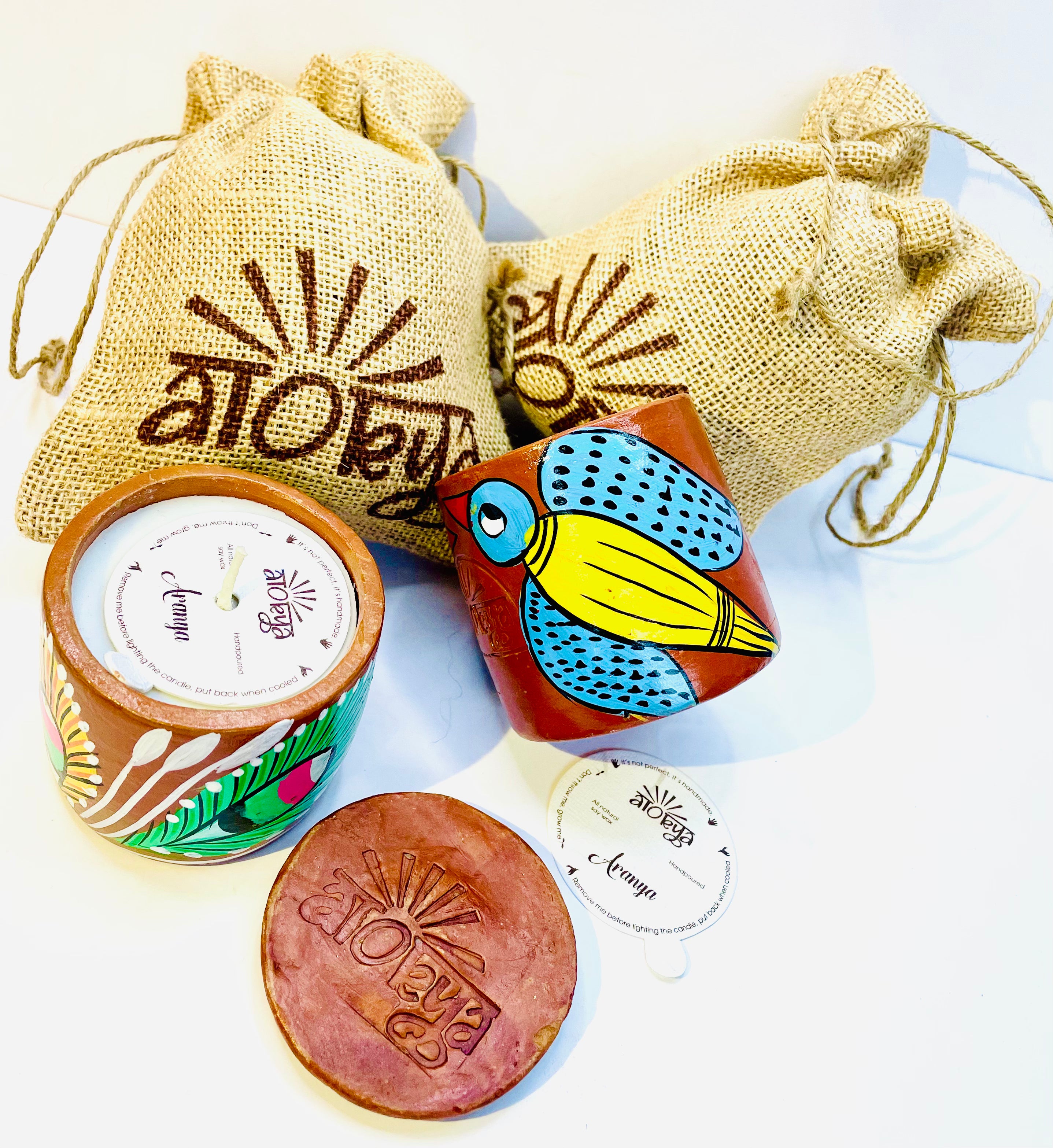 Top view of two 100% natural soy wax scented candles, one hand-painted with green and yellow feathers and the other with yellow bird having blue wings and head are placed against two jute bags with one terracotta clay candle snuffer and a seed paper displayed in between the scented candles