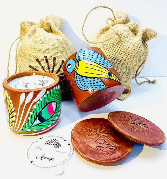 Two 100% natural soy wax scented candles, one hand-painted with green and yellow feathers and the other with yellow bird having blue wings and head are placed against two jute bags and two terracotta clay candle snuffers and seed paper dust cover is displayed in the foreground.