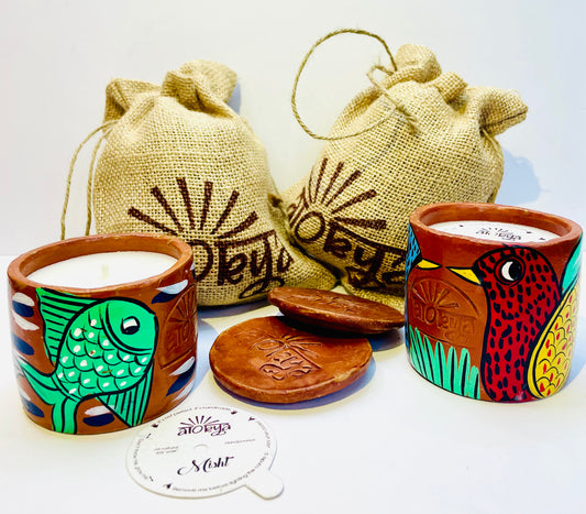 Two 100% natural soy wax scented candles, one hand-painted with green fish and the other with red bird having yellow wings and beak are placed against two jute bags with two terracotta clay candle snuffers and seed paper dust cover displayed in between.
