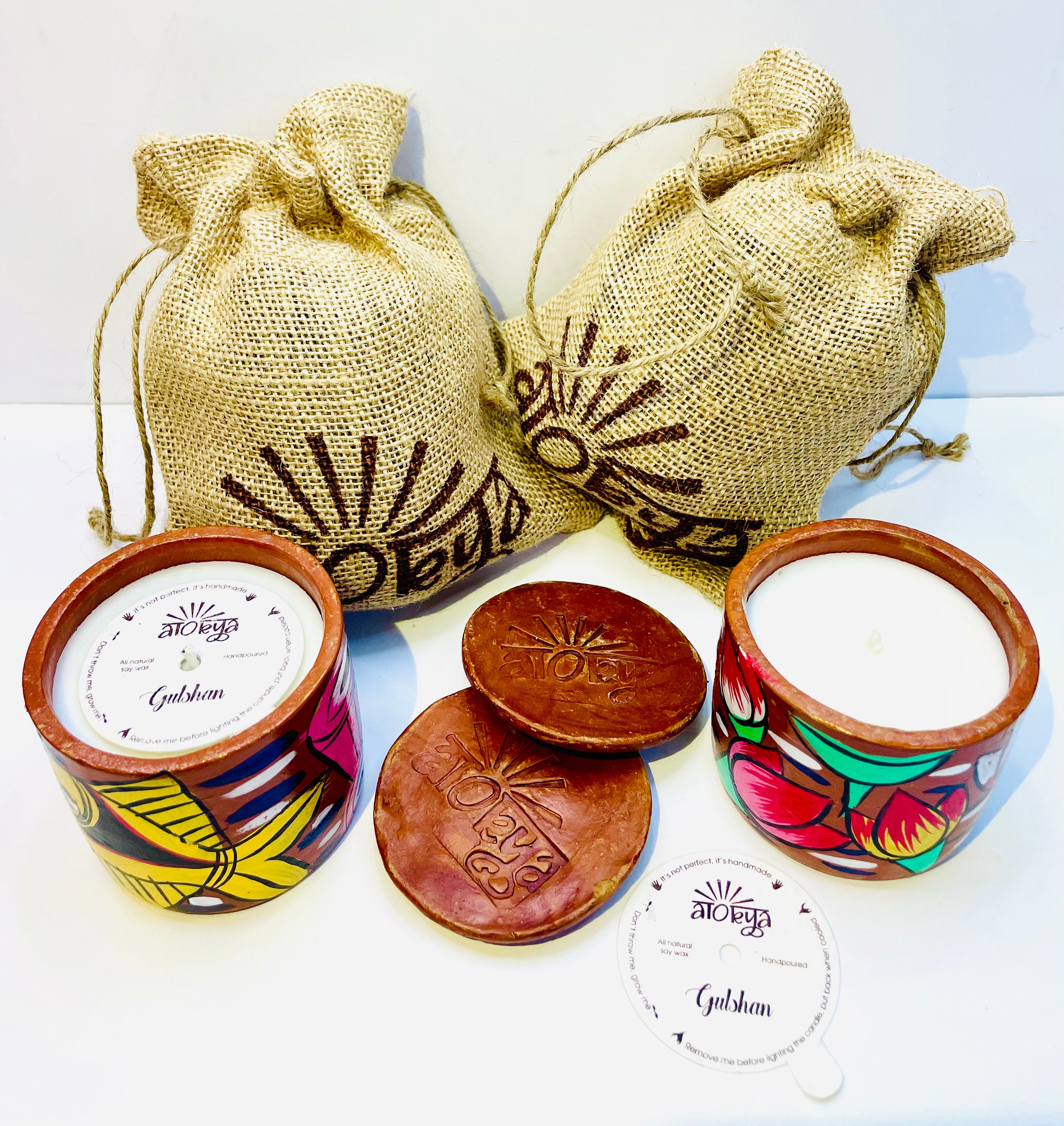 Top view of two 100% natural soy wax scented candles, one hand-painted with yellow fish and the other with pink lotus and green leaves are placed against two jute bags with two terracotta clay candle snuffers and seed paper dust covers displayed in between the scented candles