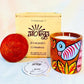 100% natural soy wax scented candle in a terracotta jar that is hand painted with a blue bird and yellow leaves motif. A seed paper candle dust cover is placed near the scented candle and has a wooden candle box and candle snuffer in the background.