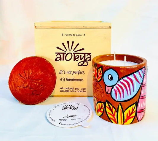 100% natural soy wax scented candle in a terracotta jar that is hand painted with a blue bird and yellow leaves motif. A seed paper candle dust cover is placed near the scented candle and has a wooden candle box and candle snuffer in the background.