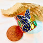 100% natural soy wax scented candle in a terracotta jar, hand painted with yellow butterfly having blue wings, is placed near terracotta clay candle snuffers and honeycomb paper.