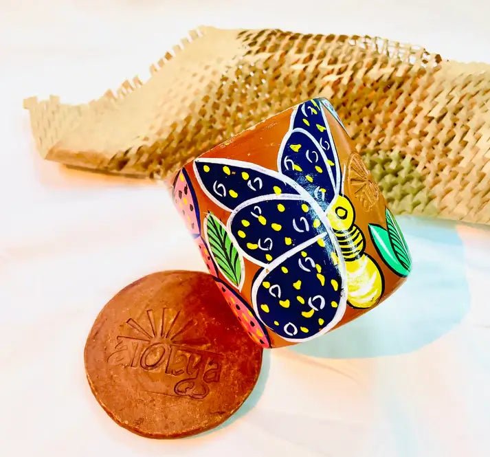 100% natural soy wax scented candle in a terracotta jar, hand painted with yellow butterfly having blue wings, is placed near terracotta clay candle snuffers and honeycomb paper.