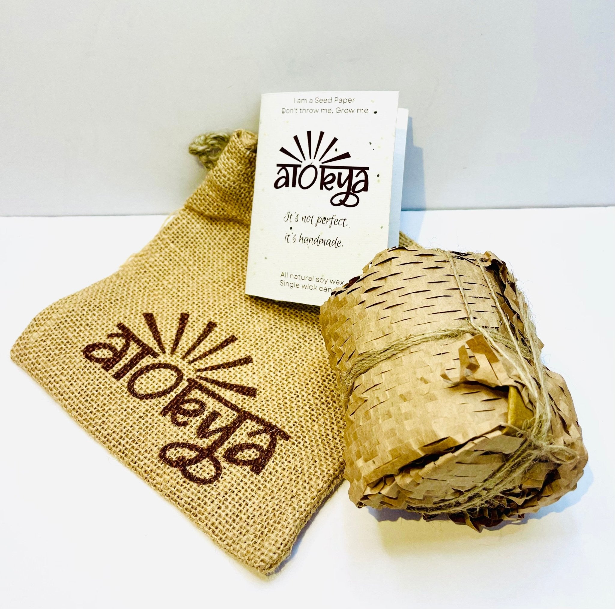  A 100% natural soy wax-scented candle wrapped in honeycomb paper is placed near a seed paper information card and jute bag.