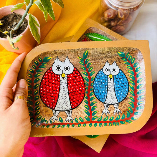 A hand holding a rectangle wood tray handcrafted from pure mango wood and featuring two Pattachitra owl motifs, hand painted by the generational Pattachitra artists of Pingla
