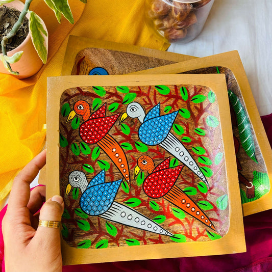 A hand holding a square wooden platter, handcrafted from pure mango wood and featuring two blue birds, two red birds and tree branches motifs, hand painted by the generational Pattachitra artists of Pingla.