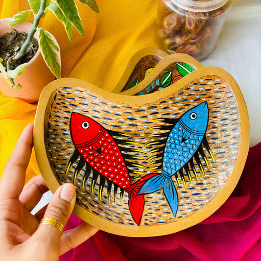 A hand holding a moon-shaped wooden platter tray handcrafted from mango wood and featuring two fish motifs, hand painted by the generational Pattachitra artists of Pingla