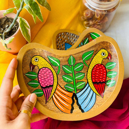 A hand holding a moon-shaped wooden platter tray or trinket tray handcrafted from mango wood and featuring a bird couple motif, hand painted by the generational Pattachitra artists of Pingla