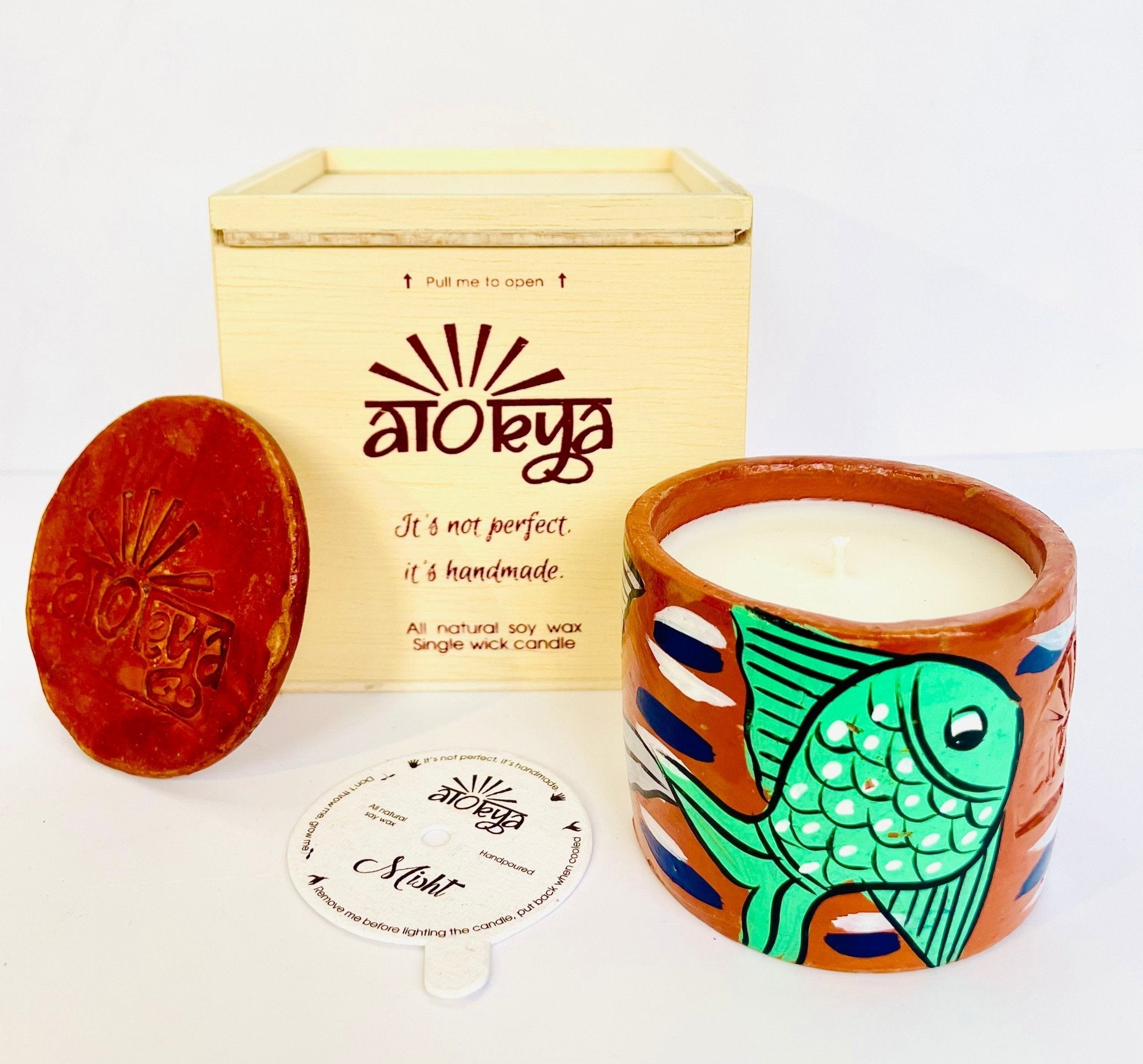 100% natural soy wax scented candle in a terracotta jar that is hand-painted with a green fish motif. A seed paper candle dust cover is placed near the scented candle and has a wooden candle box and candle snuffer in the background. 