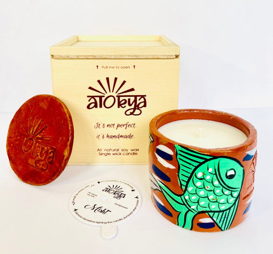 100% natural soy wax scented candle in a terracotta jar that is hand-painted with a green fish motif. A seed paper candle dust cover is placed near the scented candle and has a wooden candle box and candle snuffer in the background. 