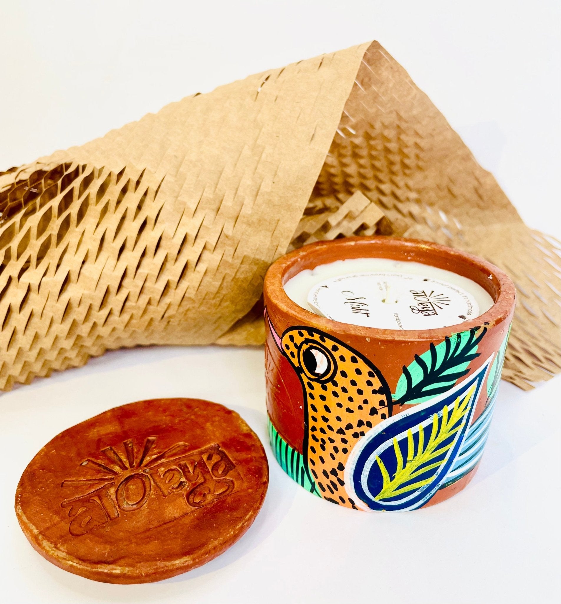 100% natural soy wax scented candle in a terracotta jar hand-painted with an orange bird having blue and yellow feathers is covered with a seed paper candle dust cover while honeycomb paper and candle snuffer is placed around the candle