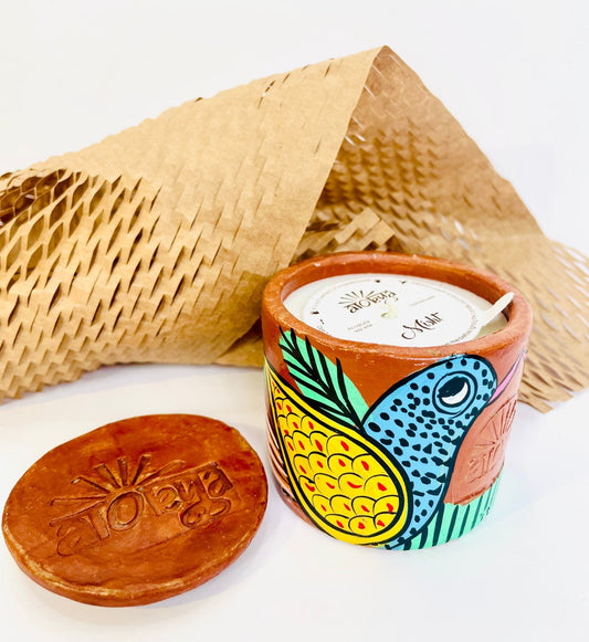 100% natural soy wax scented candle in a terracotta jar hand-painted with a blue coloured bird having yellow feathers is covered with a seed paper candle dust cover while honeycomb paper and candle snuffer are placed around the candle