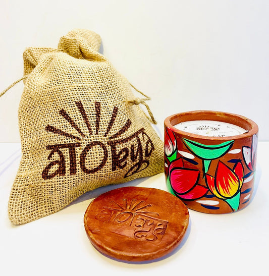 100% natural soy wax scented candle in a terracotta jar, hand-painted with one red lotus and one orange-yellow lotus surrounded by green leaves motif is covered with seed paper dust cover and a jute bag and terracotta clay candle snuffer is placed near the aromatherapy scented candle. 