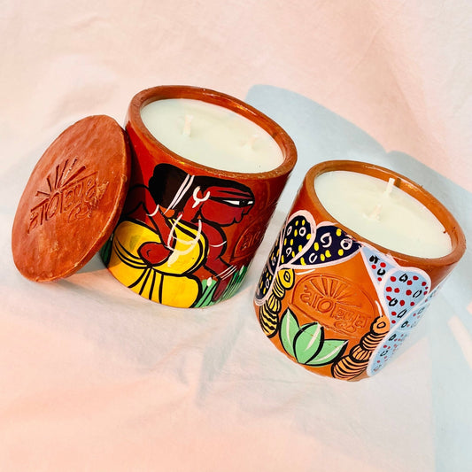 Two 100% natural soy wax scented candles, one hand-painted with a tribal woman and the other with yellow butterflies having blue wings are placed near a terracotta clay candle snuffer
