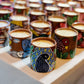 100% natural soy wax scented candles in terracotta jars with paintings of birds, peacocks and fish are displayed on a table covered with white cloth.