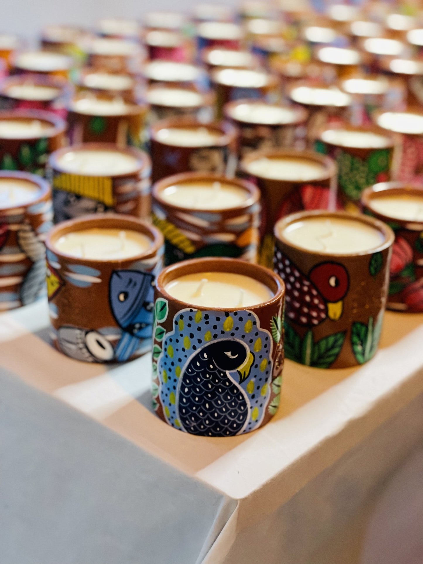 100% natural soy wax scented candles in terracotta jars with paintings of birds, peacocks and fish are displayed on a table covered with white cloth.